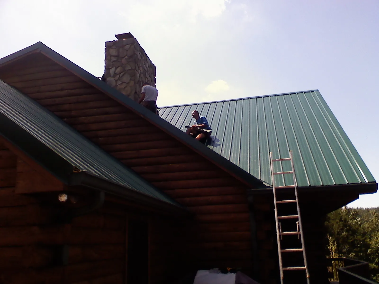 Roofers installing green metal panels beside a stone chimney on a log home in Orangeburg